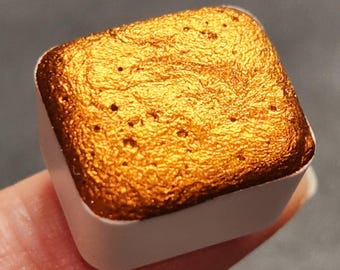 May include: A square pan of metallic gold watercolor paint. The paint is in a white pan with a smooth, metallic finish.