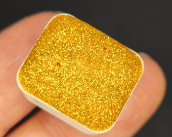 Puede incluir: Un pequeño recipiente cuadrado lleno de pintura dorada con purpurina. La pintura es de un color dorado metálico brillante y tiene una textura fina y brillante.