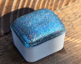 May include: A small square container with a lid. The lid is covered in a sparkly blue glitter with iridescent flecks. The container is white.