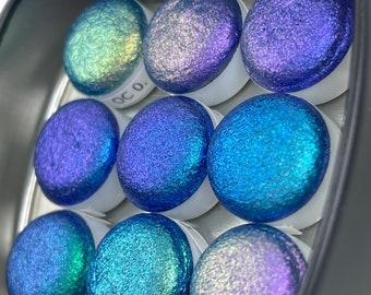 May include: A set of nine round, iridescent paint samples in a metal tin. The samples are arranged in a circle and have a variety of blue, green, and purple hues.