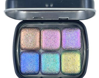 Puede incluir: Una pequeña caja metálica azul oscuro abierta que contiene seis pastillas de acuarela cuadradas. Las pastillas están llenas de colores brillantes e iridiscentes, incluyendo rosa, morado, azul y amarillo. La caja está abierta, revelando las pinturas.