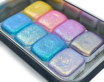 May include: A set of eight iridescent watercolor paints in a metal tin. The colors are yellow, pink, blue, purple, and green. The paint pans are labeled with the names "Ruri" and "Hana".