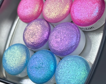 May include: A tin with eight round iridescent paint pots. The paint pots are in shades of pink, purple, blue and green. The tin has the word "Mulan" printed on it.