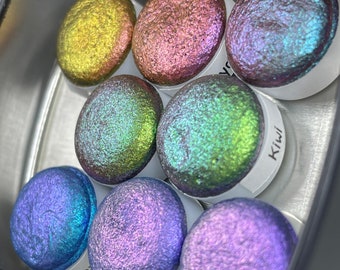 May include: A set of seven iridescent watercolour paints in a metal tin. The colours are green, blue, purple, pink, and gold.