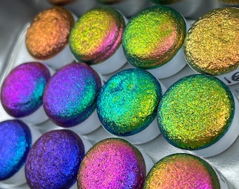 May include: A collection of iridescent paint samples in a variety of colors, including green, blue, purple, pink, and orange. The samples are arranged in a circular pattern in a metal tin.