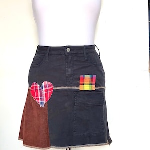 Peut inclure: Jupe en jean gris foncé avec un panneau en velours côtelé marron et une application en forme de cœur à carreaux rouges. Une petite poche présente un patch à carreaux colorés. La jupe est présentée sur un mannequin avec un haut tube blanc.