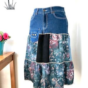 May include: A denim skirt with a patchwork design. The top section is classic blue denim, while the lower part features floral print panels in shades of blue, green, and red. A black panel is centered on the front.