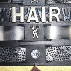 May include: A black leather bracelet with multiple charms, including a silver shield with a design, a red lip, a skull and crossbones, the word "HAIR", a pair of scissors, a clear rhinestone, a gold diamond, a purple leather band with a rhinestone circle, and a blue leather band with a green rhinestone.