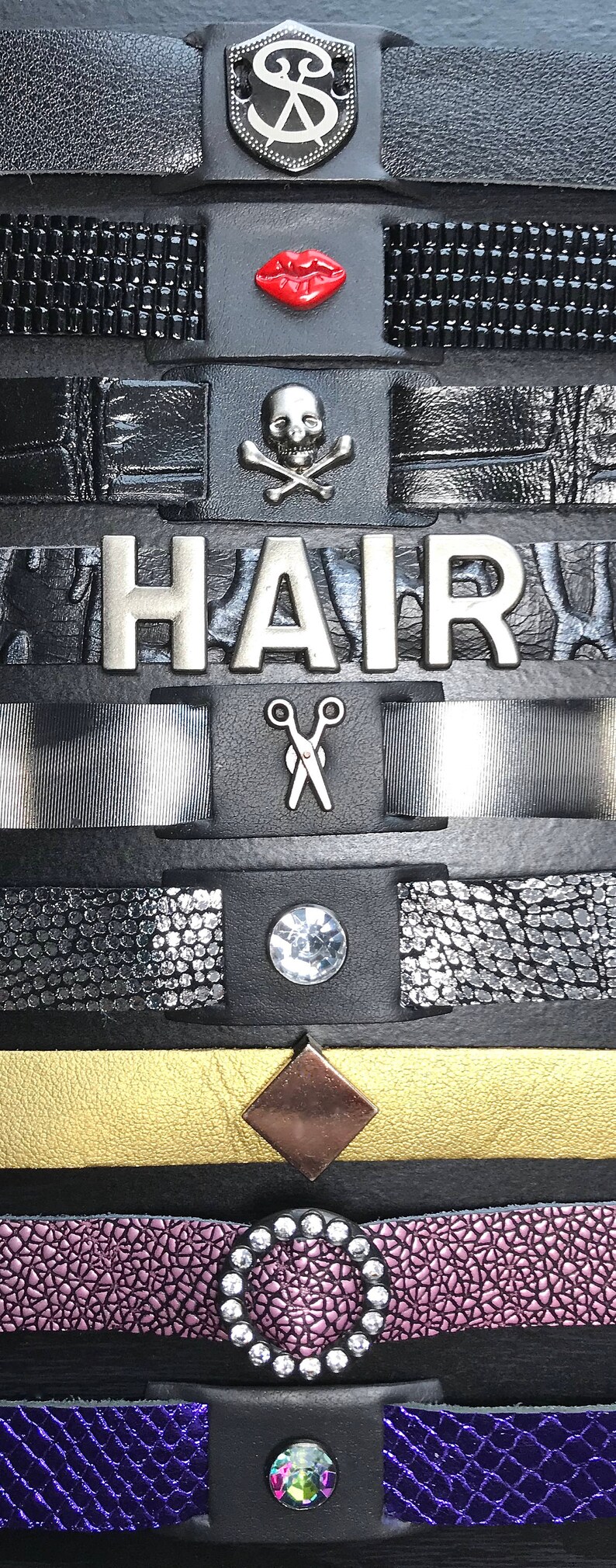 May include: A black leather bracelet with multiple charms, including a silver shield with a design, a red lip, a skull and crossbones, the word "HAIR", a pair of scissors, a clear rhinestone, a gold diamond, a purple leather band with a rhinestone circle, and a blue leather band with a green rhinestone.