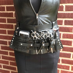 May include: Black leather belt with multiple pockets and a crocodile-embossed design. The belt has a phone holder, multiple slots for scissors, and a small pocket for other tools.