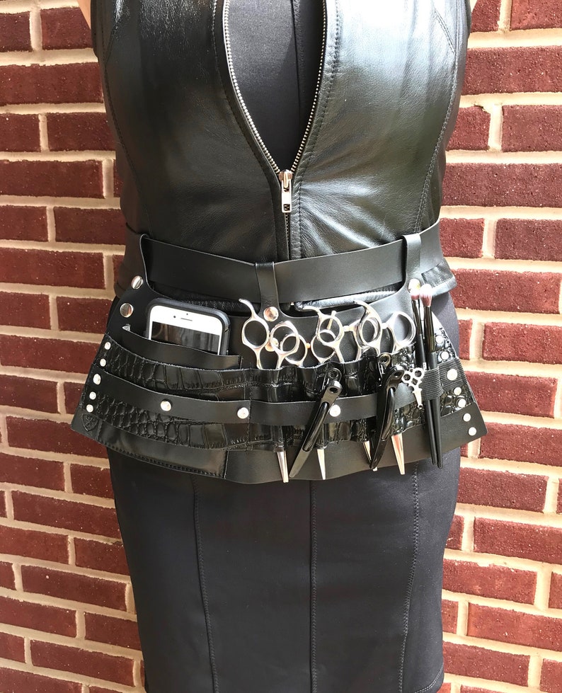 May include: Black leather belt with multiple pockets and a crocodile-embossed design. The belt has a phone holder, multiple slots for scissors, and a small pocket for other tools.