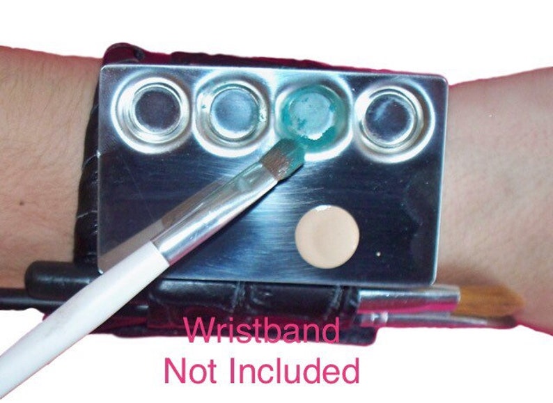 Puede incluir: Una paleta de maquillaje de plata con cinco pozos circulares, uno lleno de una sustancia verde claro, otro de una sustancia marr&oacute;n claro y los otros tres vac&iacute;os. Una brocha de maquillaje blanca descansa sobre la paleta. La paleta est&aacute; unida a una pulsera de cuero negro. El texto "Wristband Not Included" est&aacute; impreso debajo de la paleta.