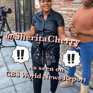 May include: A woman wearing a black leather shirt and black pants with a tool belt. She is standing in front of a brick wall with a camera and tripod in the background. The text "@SheritaCherry" and "As seen on CBS World News Report" is visible in the image.