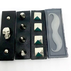 Puede incluir: Cuatro parches de cuero negro con diferentes adornos. El primer parche tiene una calavera plateada, el segundo tiene tres pinchos plateados, el tercero tiene cuatro tachuelas piramidales plateadas y el cuarto tiene un bigote de fieltro gris.