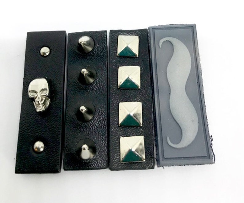 Puede incluir: Cuatro parches de cuero negro con diferentes adornos. El primer parche tiene una calavera plateada, el segundo tiene tres pinchos plateados, el tercero tiene cuatro tachuelas piramidales plateadas y el cuarto tiene un bigote de fieltro gris.