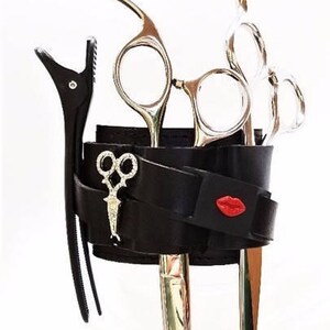 May include: Black leather wrist strap with a red lip design, holding a set of silver hairdressing shears and a silver hair clip.
