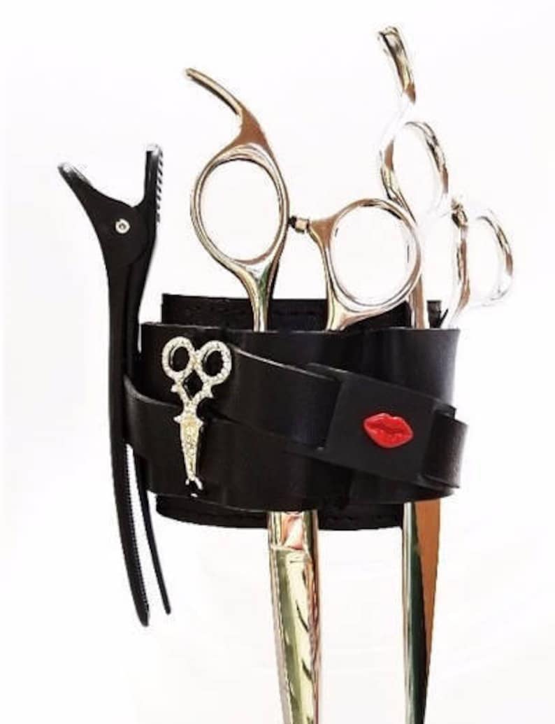 May include: Black leather wrist strap with a red lip design, holding a set of silver hairdressing shears and a silver hair clip.