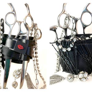 Könnte beinhalten: Schwarzer Leder-Scherenhalter mit silbernen Ketten und Strasssteinen. Der Halter hat zwei Schlitze für Scheren und eine kleine Tasche. Auf der Vorderseite des Halters befindet sich ein rotes Lippenmotiv.