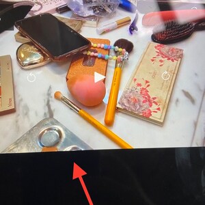Puede incluir: Primer plano de un tocador de maquillaje con varios art&iacute;culos, incluyendo un tel&eacute;fono inteligente negro, un estuche dorado, una esponja de maquillaje rosa, una brocha de maquillaje amarilla, una paleta de maquillaje plateada y una paleta de maquillaje con estampado floral con la palabra "Jouer" impresa en ella.