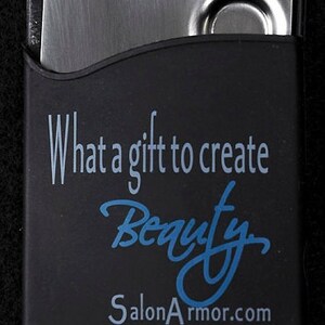 Puede incluir: Una funda de silicona negra con un inserto de metal plateado. La funda tiene el texto "What a gift to create Beauty SalonArmor.com" impreso en ella.
