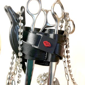 May include: Black leather scissor holder with a red lip design. The holder has two pairs of silver scissors and a silver chain with rhinestones.