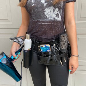May include: A black leather stylist belt with multiple pockets and loops for holding tools. The belt has a phone holder, a comb holder, a scissor holder, and a bottle holder. The belt is worn over black pants and a grey t-shirt with the text "The Breakfast Club".