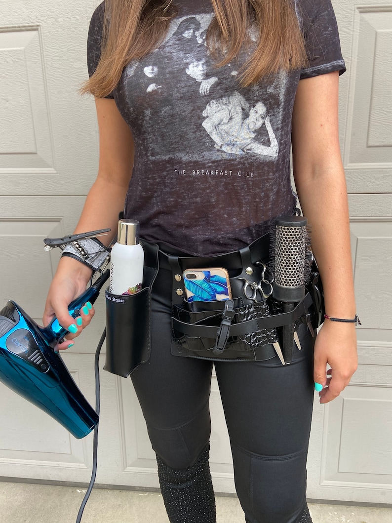May include: A black leather stylist belt with multiple pockets and loops for holding tools. The belt has a phone holder, a comb holder, a scissor holder, and a bottle holder. The belt is worn over black pants and a grey t-shirt with the text "The Breakfast Club".