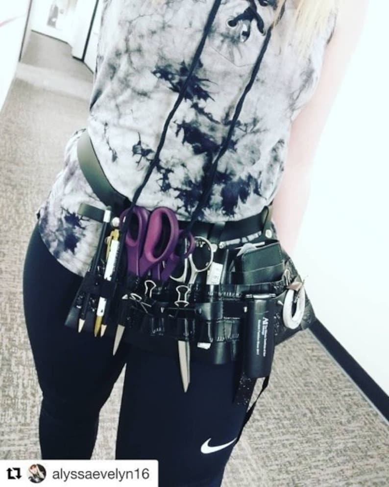 May include: A person wearing a black and white tie-dye tank top with a black belt and a black utility belt with various tools and accessories, including scissors, pens, and a small spray bottle. The person is wearing black leggings with a white Nike swoosh logo on the left leg.