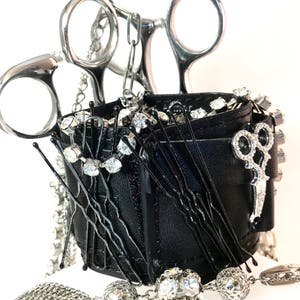 May include: A black leather wristlet with a silver chain and a pair of silver scissors. The wristlet is filled with black hair pins and clear rhinestones.