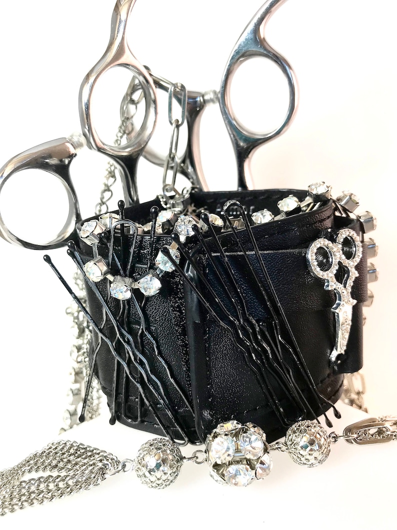 May include: A black leather wristlet with a silver chain and a pair of silver scissors. The wristlet is filled with black hair pins and clear rhinestones.