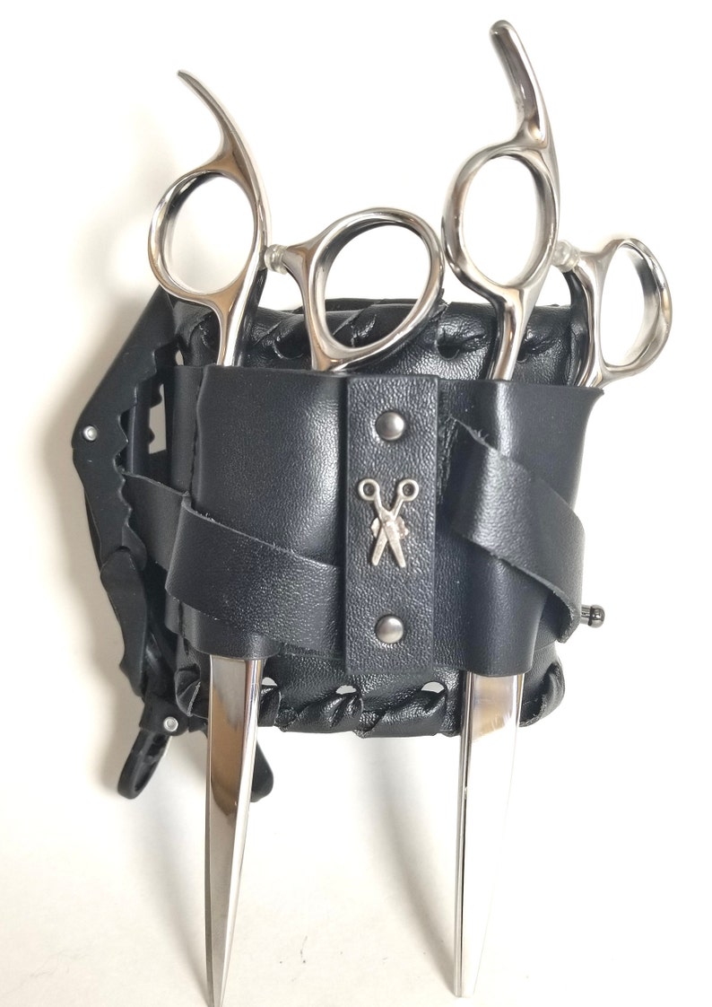 Puede incluir: Una correa de mu&ntilde;eca de cuero negro con un dise&ntilde;o trenzado sostiene tres tijeras de peluquer&iacute;a plateadas. La correa tiene un logotipo de tijeras plateadas y se asegura con un cierre a presi&oacute;n.