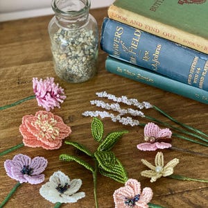 May include: Beaded flowers in shades of pink, white, and lavender, with green stems, are arranged on a wooden surface. A glass jar filled with small stones and a stack of vintage books, including "Butterflies & Moths", are also visible.