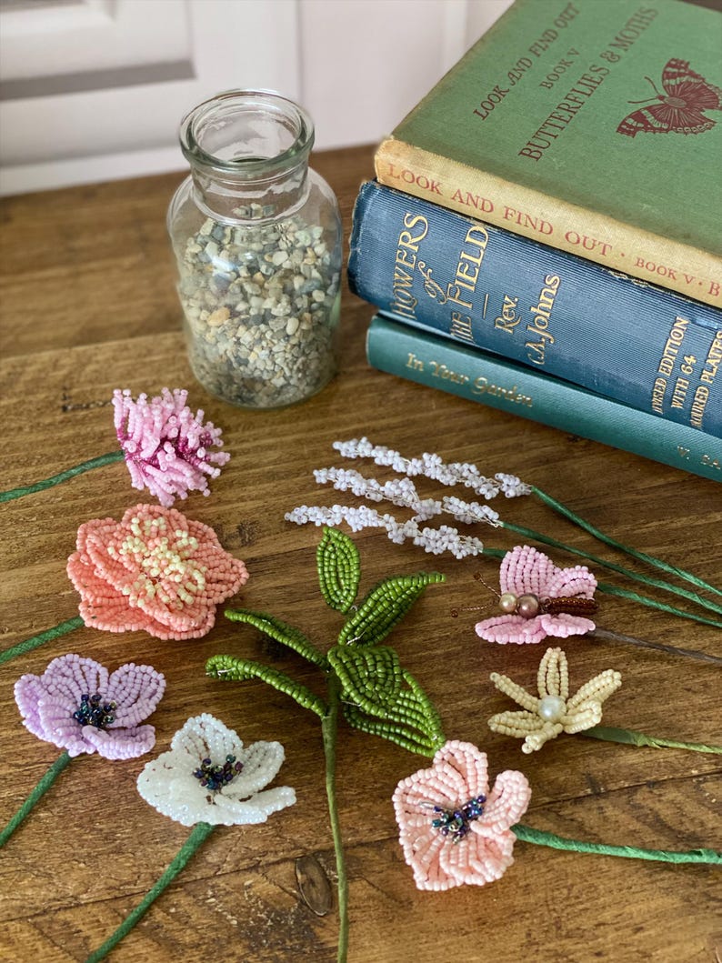 May include: Beaded flowers in shades of pink, white, and lavender, with green stems, are arranged on a wooden surface. A glass jar filled with small stones and a stack of vintage books, including "Butterflies & Moths", are also visible.