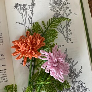 May include: An open book displays botanical illustrations with two beaded flowers. One flower is coral, the other is pink. Green plastic fern-like leaves and stems are also visible.