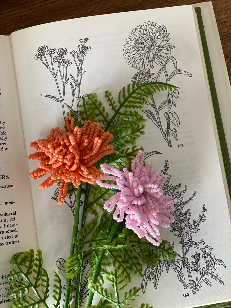 May include: An open book displays botanical illustrations with two beaded flowers. One flower is coral, the other is pink. Green plastic fern-like leaves and stems are also visible.