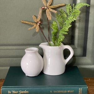 May include: A still life arrangement with a white jug and vase, adorned with beaded dragonfly ornaments and greenery. The items rest on a stack of green books with gold lettering, including "In Your Garden".