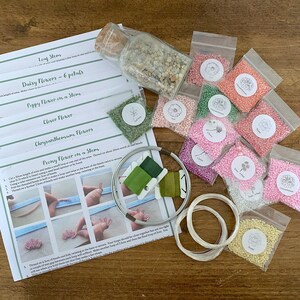 May include: A craft kit for creating beaded flowers, featuring instructions and materials. The kit contains small bags of coloured beads, green thread, wire, and a glass jar of small stones. Instructions for leaf, daisy, poppy, clover, chrysanthemum, and peony flowers are included.