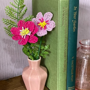 May include: A small pink vase displays beaded flowers in pink and yellow, with green leaves. Two green books with gold lettering are positioned behind the vase. A small pink glass jar is also visible.