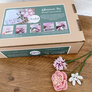 May include: A craft kit for creating beaded flowers, presented in a box labelled "Afternoon Tea Beaded Vase Craft Kit." The kit contains beads, wire, and instructions. Three finished beaded flowers in pink, white, and purple are shown on a wooden surface.