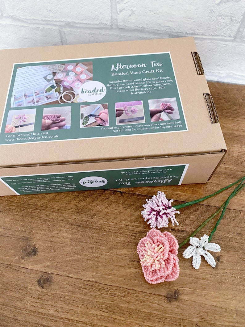 May include: A craft kit for creating beaded flowers, presented in a box labelled "Afternoon Tea Beaded Vase Craft Kit." The kit contains beads, wire, and instructions. Three finished beaded flowers in pink, white, and purple are shown on a wooden surface.