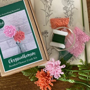 May include: A craft kit for making beaded chrysanthemum flowers. The kit includes a box with the words "Chrysanthemum Beaded Flower Craft Kit", beads in pink and orange, green thread, and a book with botanical illustrations. Two finished flowers are also visible.