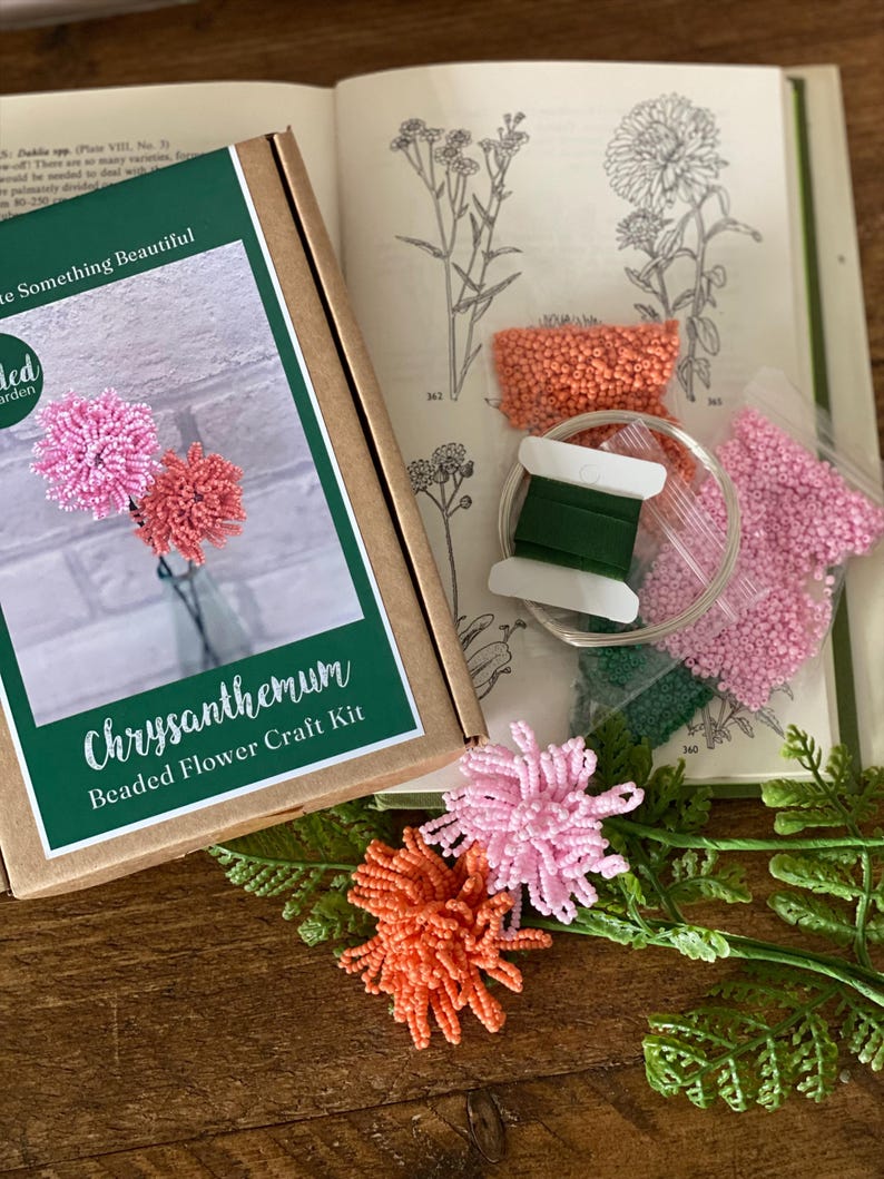 May include: A craft kit for making beaded chrysanthemum flowers. The kit includes a box with the words "Chrysanthemum Beaded Flower Craft Kit", beads in pink and orange, green thread, and a book with botanical illustrations. Two finished flowers are also visible.