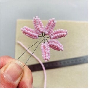 May include: A handmade pink beaded flower hair accessory. The flower has six petals and is attached to a wire stem. The accessory is held by a person's fingers, with a beige background and a ruler visible.
