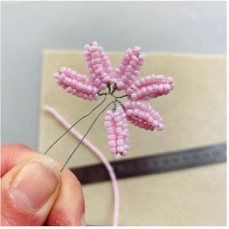 May include: A handmade pink beaded flower hair accessory. The flower has six petals and is attached to a wire stem. The accessory is held by a person's fingers, with a beige background and a ruler visible.