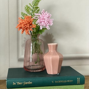 May include: A pink glass vase with beaded orange and pink flowers and green fern. A small, pink, geometric vase sits on a stack of two green books. The books are titled "In Your Garden" and "The Illustrated Guide to Garden Flowers."