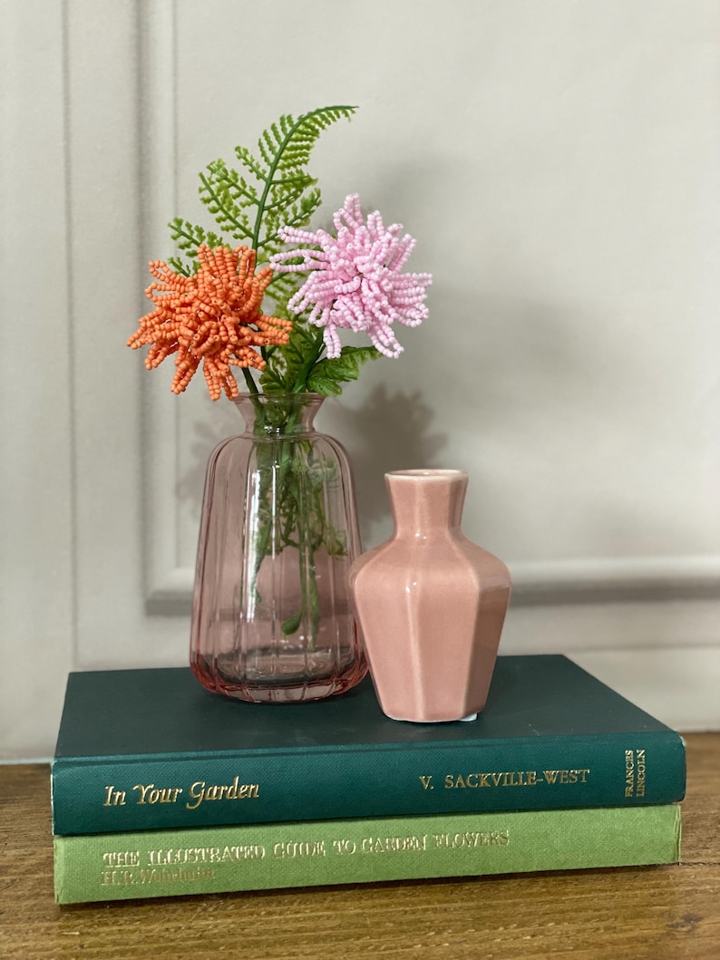 May include: A pink glass vase with beaded orange and pink flowers and green fern. A small, pink, geometric vase sits on a stack of two green books. The books are titled "In Your Garden" and "The Illustrated Guide to Garden Flowers."
