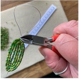 May include: A person's hand using red-handled wire cutters to trim a silver wire attached to a beaded green leaf design. A ruler and a pile of green beads are in the background. The image shows a craft project in progress.