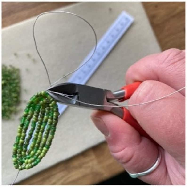 May include: A person's hand using red-handled wire cutters to trim a silver wire attached to a beaded green leaf design. A ruler and a pile of green beads are in the background. The image shows a craft project in progress.