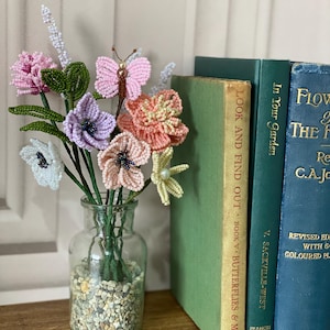 May include: A clear glass vase with a beaded flower arrangement. The flowers are in shades of pink, purple, peach, and white, with green stems and leaves. A beaded butterfly is also included. Books with green and blue covers are in the background.