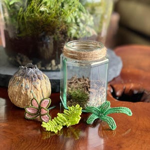 May include: A glass terrarium with greenery and a small glass jar with twine. Decorative beaded plant accents in pink, green, and a brown, textured object are arranged on a wooden surface. The image showcases a nature-inspired theme.